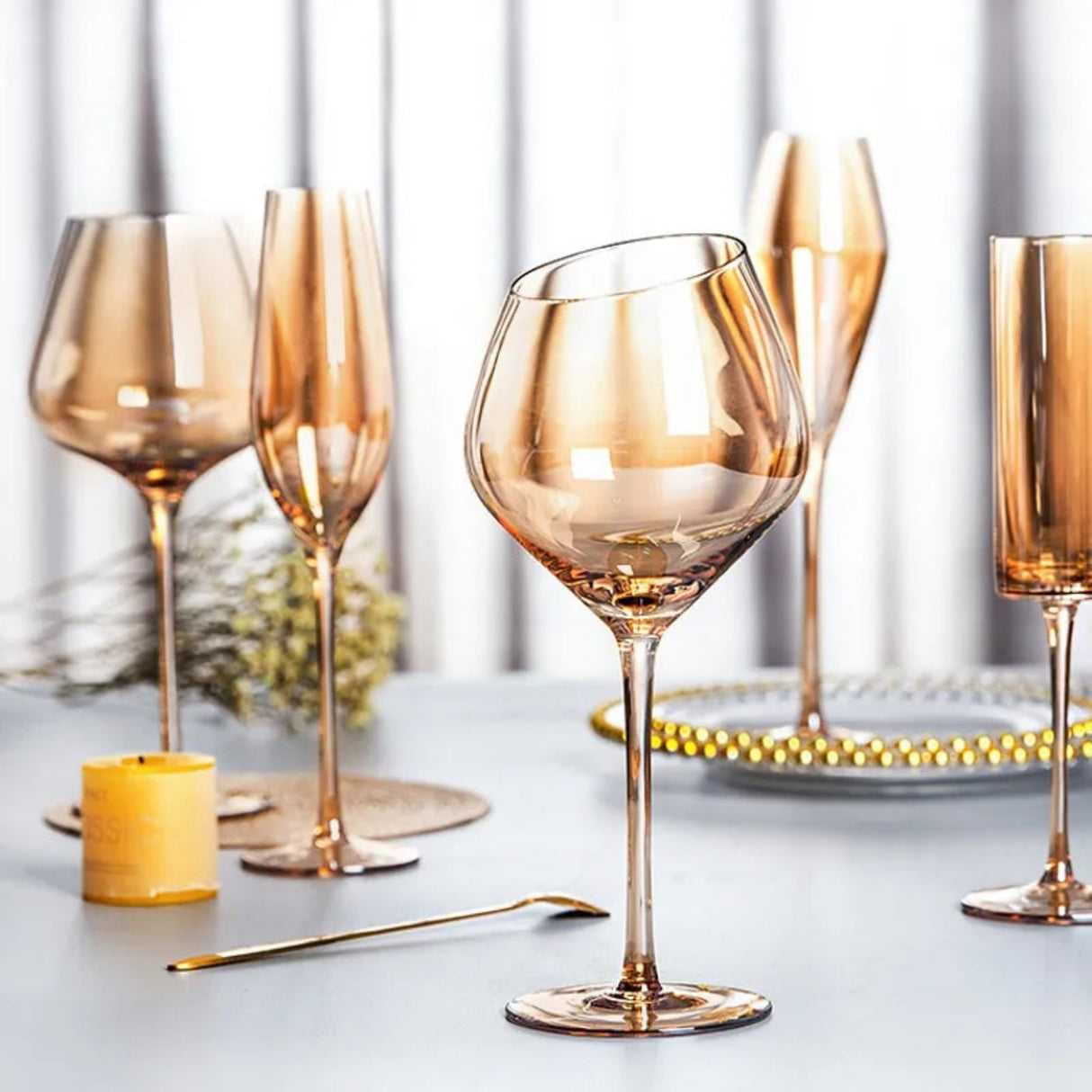 A set of elegant smoked amber glass tableware, including a wide wine glass, a champagne flute, a tumbler, and a small candle, arranged on a light gray tabletop with a gold spoon and decorative plates in the background.