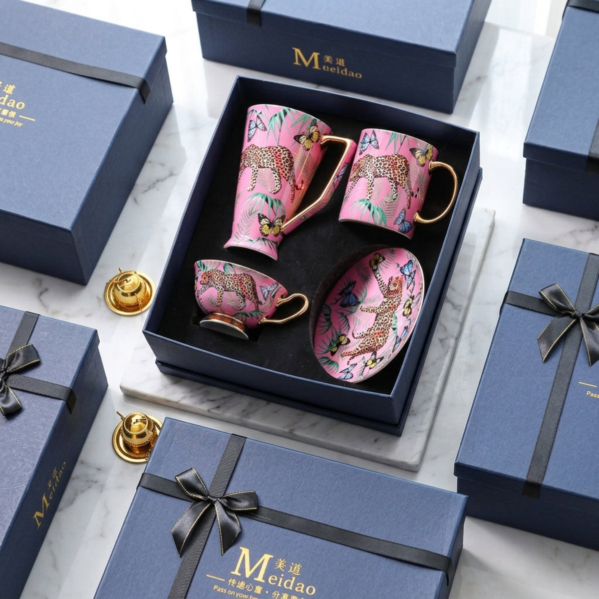 A pink ceramic mug, matching teacup, saucer, and small gold-toned accessory set arranged in an open dark blue gift box. The cups and saucer are decorated with leopard prints and multicolored floral motifs.