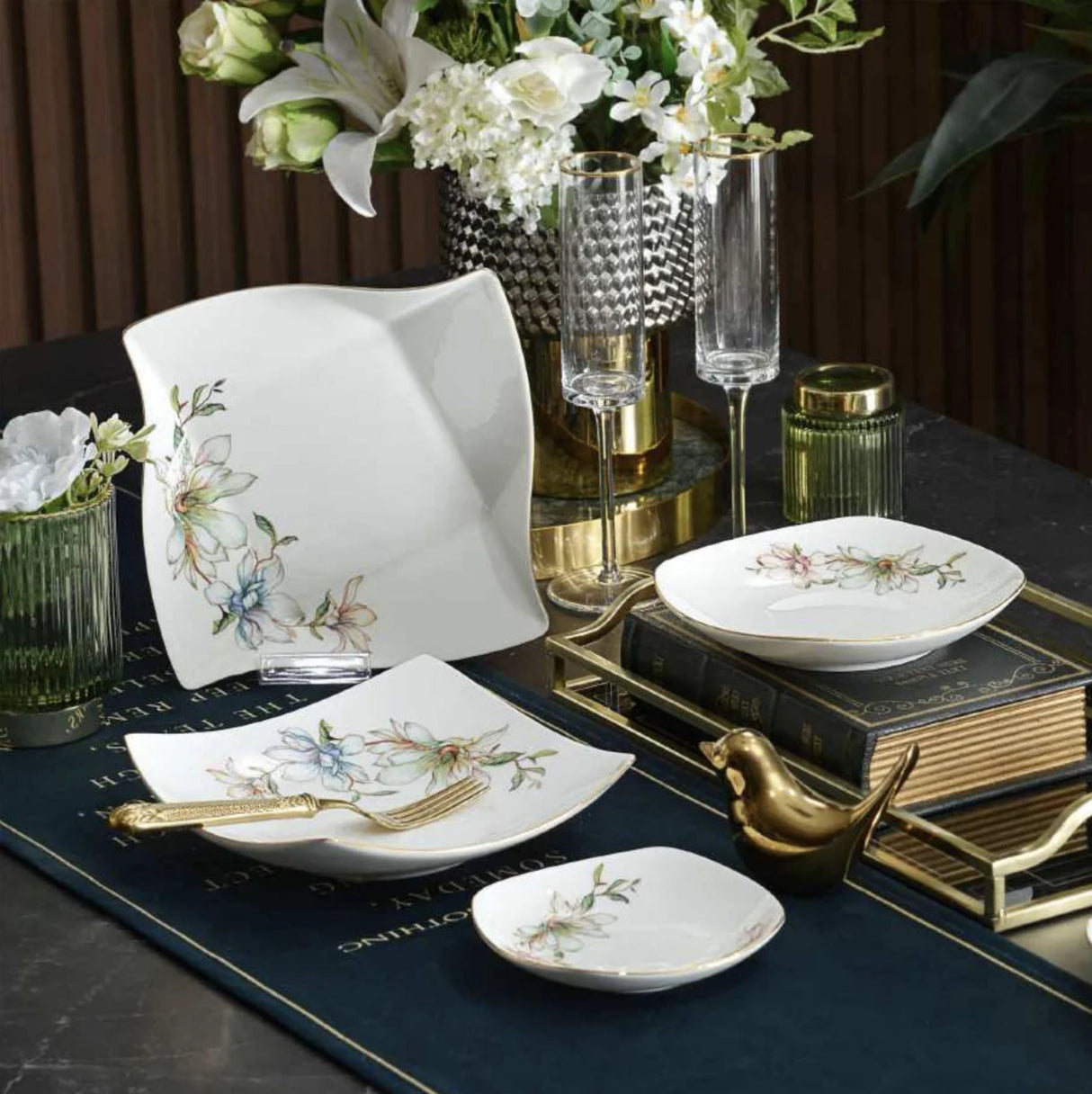 A set of white ceramic tableware with delicate floral and bird illustrations, styled on a dark table runner with gold accents, books, and glassware.