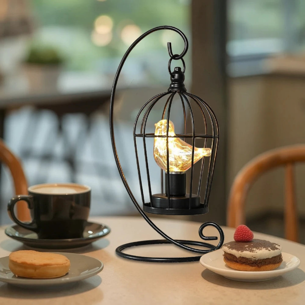 Decorative lamp with a lit bulb in a cage on a table with a cup of coffee and dessert.