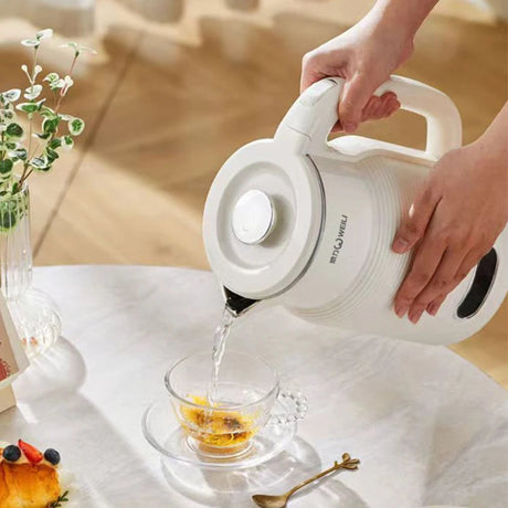 Person pouring tea from a white electric kettle into a glass cup on a white tablecloth.