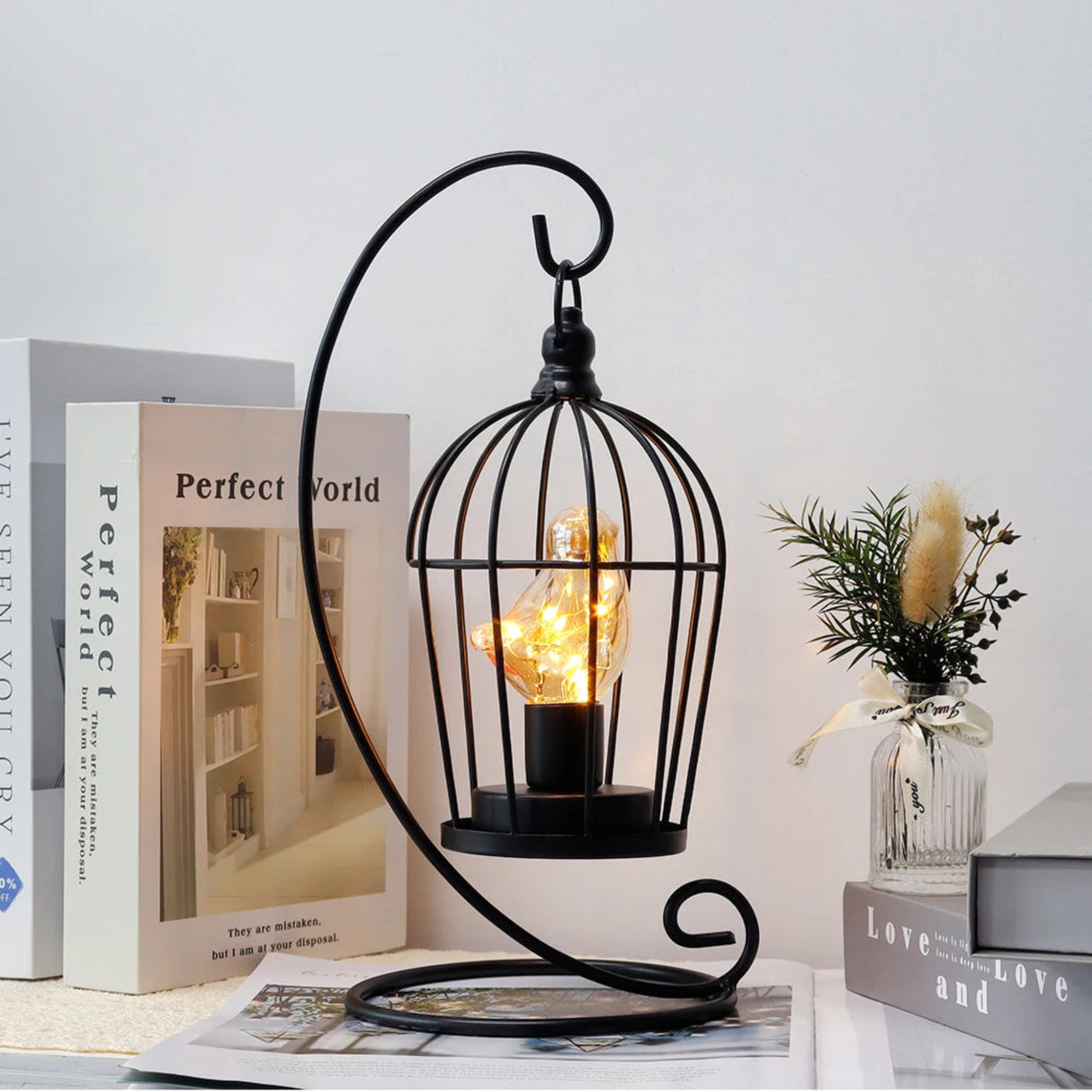 Decorative black metal lamp with a light bulb inside, placed on a surface with books and a vase in the background.