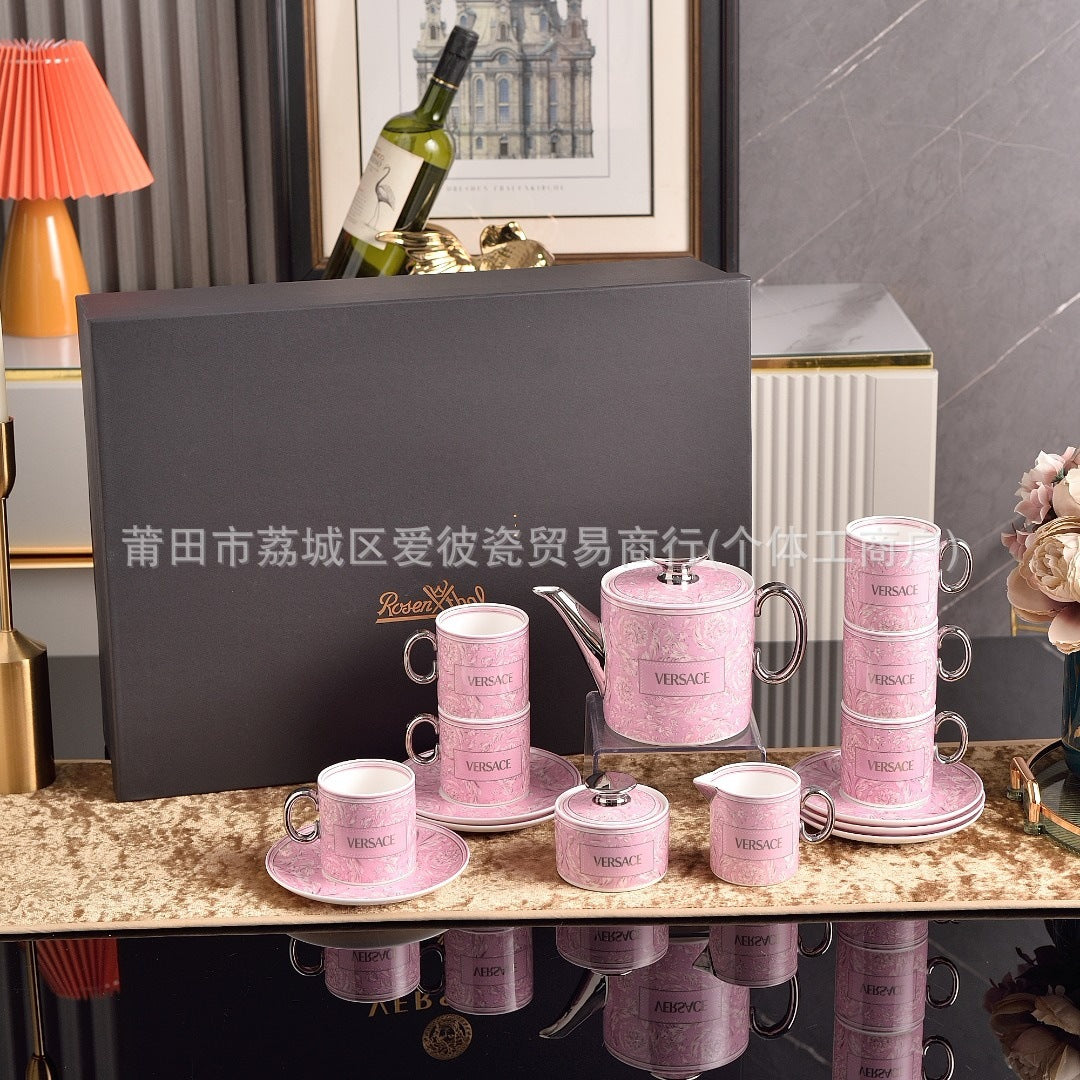A pink Versace teacup and teapot set arranged on a marble table in a styled interior, with a large black presentation box behind it. The cups, saucers, teapot, and small containers are decorated with a light pink damask pattern and the Versace logo, while the background includes flowers, a lamp, framed art, and a sofa.