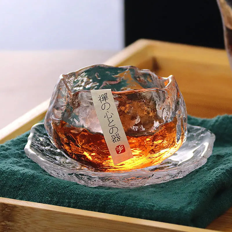 A single clear glass teacup with a brown liquid inside, placed on a wooden surface, with a green cloth underneath. The teacup has Japanese characters on it.