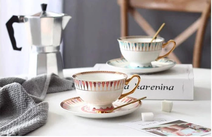 Cup and saucer set with a spoon on a table with a coffee maker in the background.