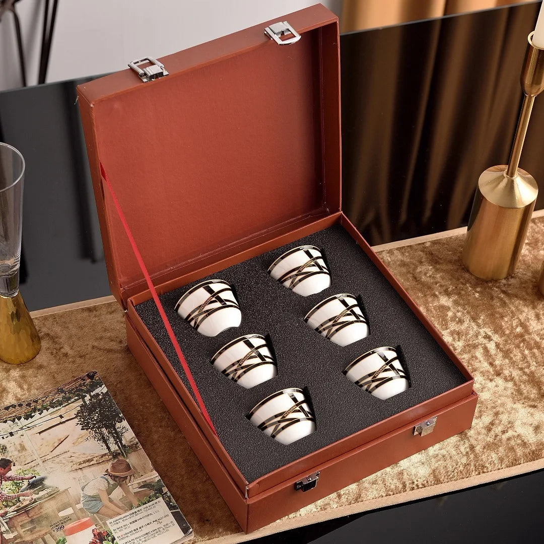 Set of four silver and white ceramic cups in a brown leather case on a decorative surface.