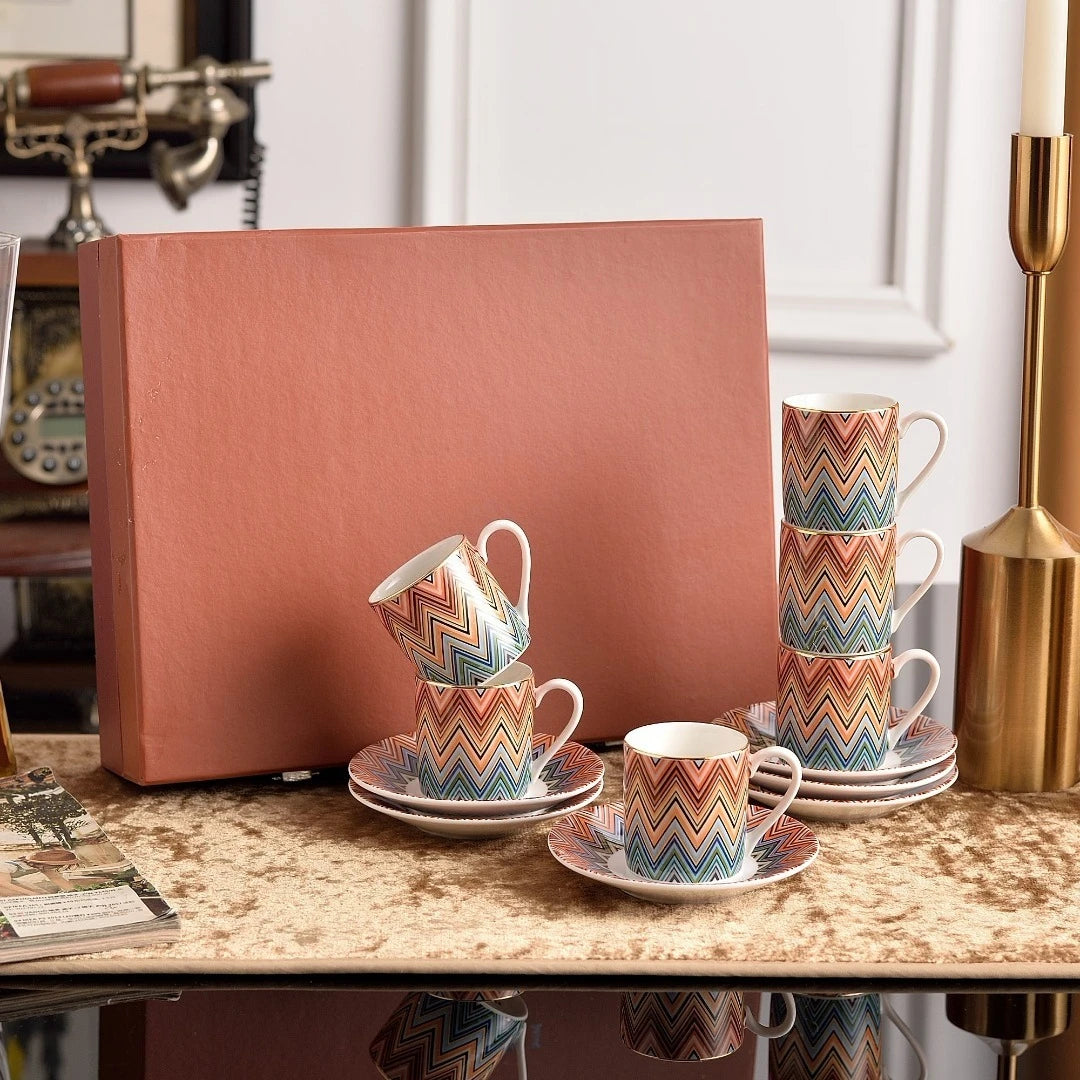 Set of colorful zigzag patterned cups and saucers on a marble surface with a pink box in the background.