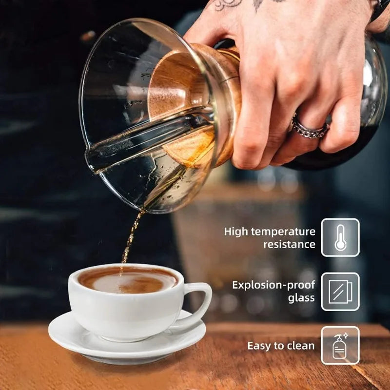 Hand pouring coffee from a glass and wood coffee maker into a white cup on a wooden table.