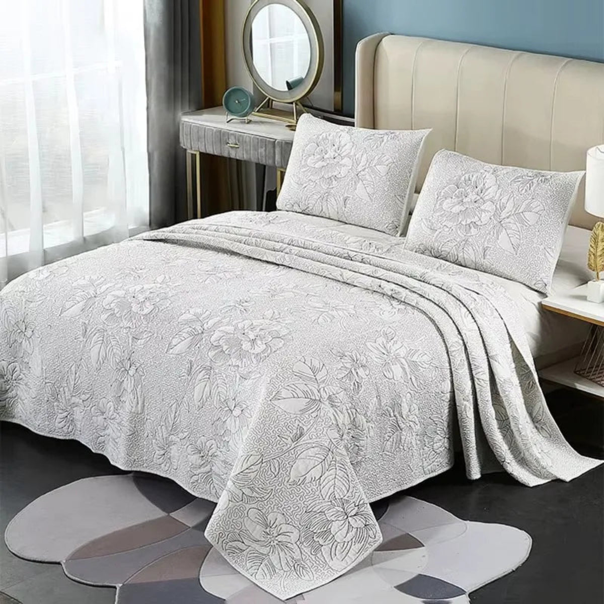 A light gray quilted bedspread with a matching floral jacquard pattern and two matching pillowcases, styled on a bed with a beige headboard in a modern bedroom setting.