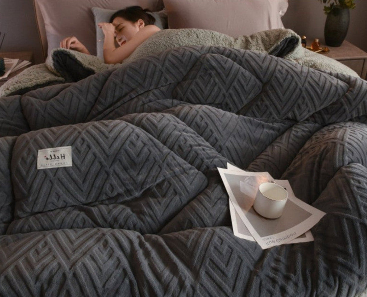 Person lying in bed under a large gray comforter with a cup and newspaper on the nightstand.