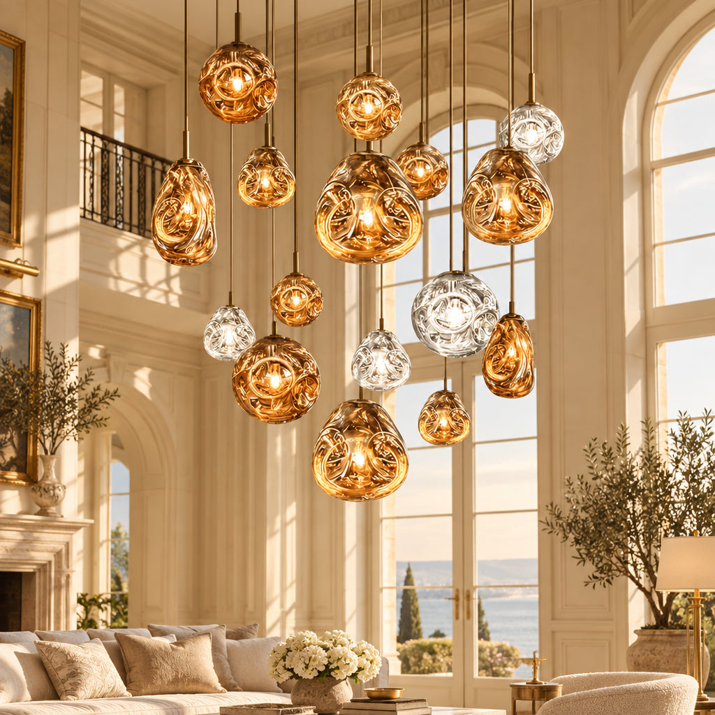 A cluster of modern glass pendant lights in amber, gold, and clear finishes hanging in a large interior space with floor-to-ceiling windows overlooking a sunset over the ocean.