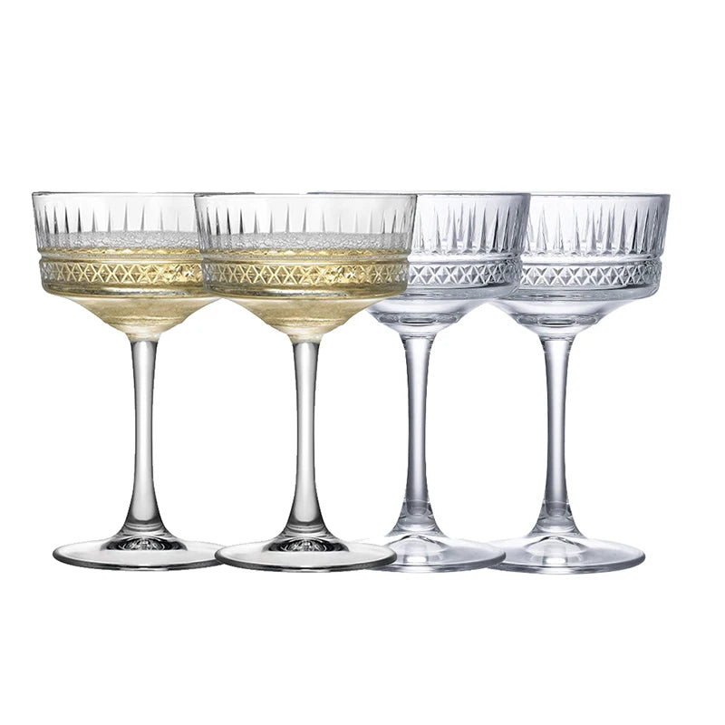 Four elegant clear coupe glasses filled with colorful cocktails arranged on a marble table in a bright indoor setting. The drinks are pink, lavender, pale yellow, and light green, garnished with citrus slices, berries, lavender sprigs, and mint leaves, with flowers and lemons in the background.