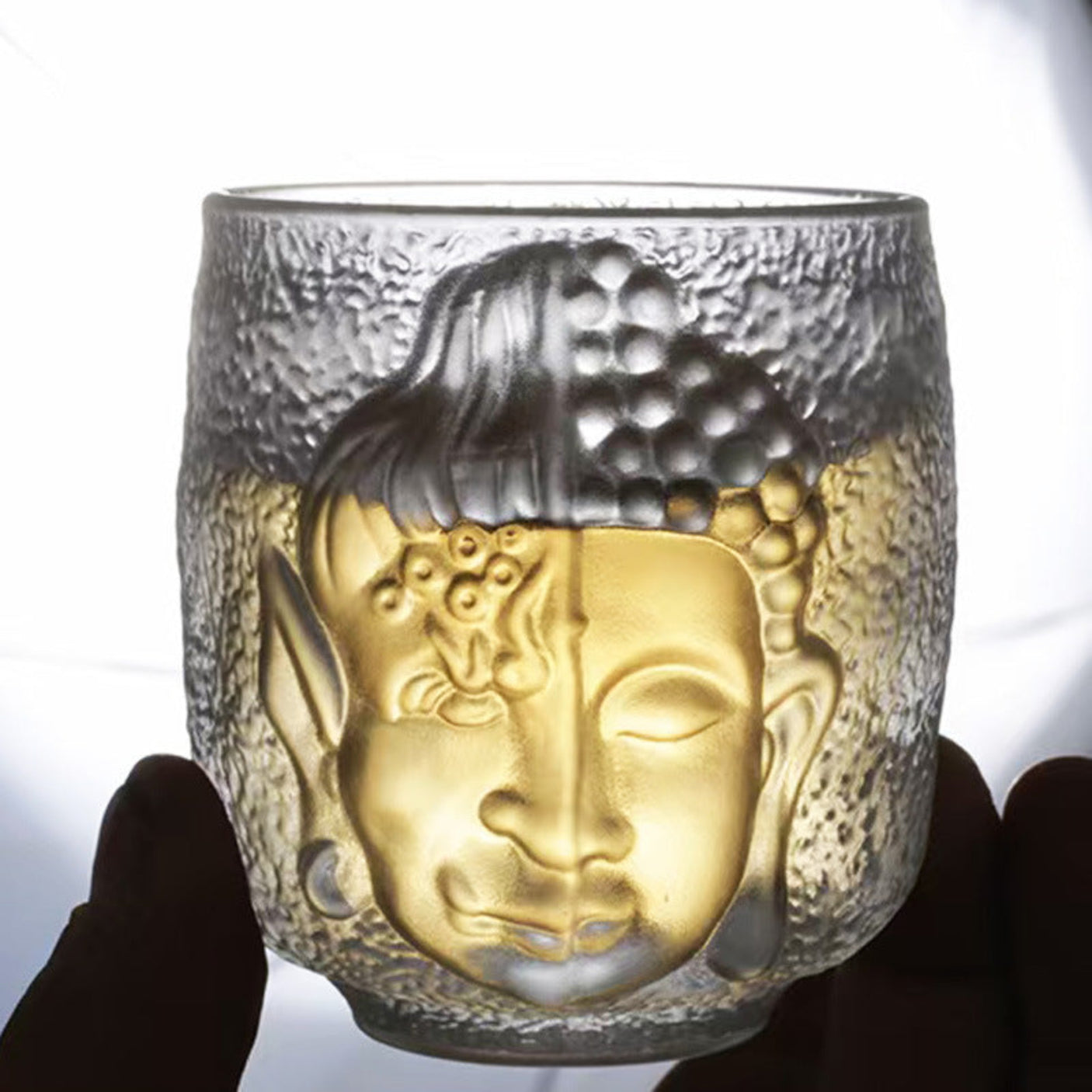 Glass tumbler with Buddha face design held by a hand against a white background