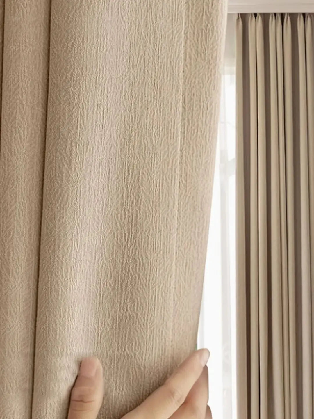 Close-up of beige curtains with a hand touching one, with light filtering through.
