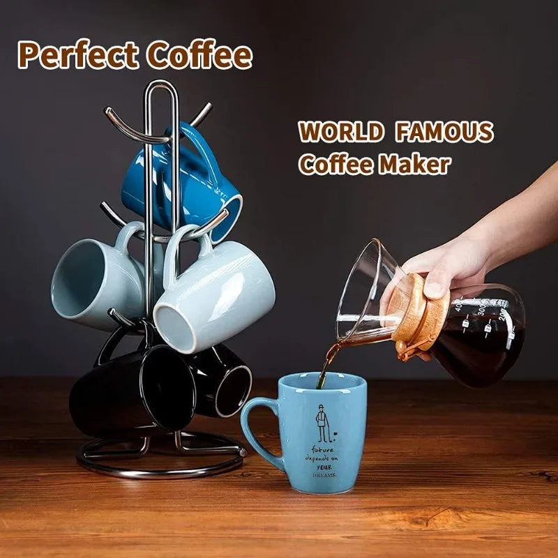 Coffee maker with mugs and a person pouring coffee into a blue mug on a wooden surface.