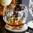 A faceted clear whiskey glass filled with amber liquor, resting on a round gray coaster printed with the word "DRINK WHISKY." Bottles of whiskey are blurred in the background, and part of another coaster and glass are visible at the edges.