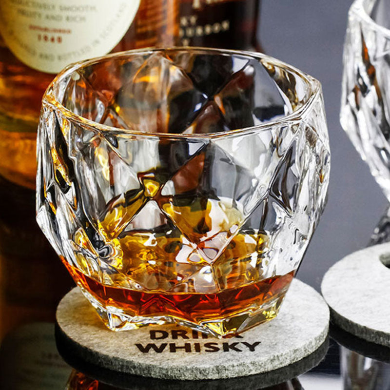 A faceted clear whiskey glass filled with amber liquor, resting on a round gray coaster printed with the word "DRINK WHISKY." Bottles of whiskey are blurred in the background, and part of another coaster and glass are visible at the edges.