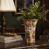 A tall, flared ceramic vase with a mottled cream, rust-orange, and green glaze, filled with green foliage, displayed on a dark marble side table in a classic, elegant room next to a brass lamp and books.