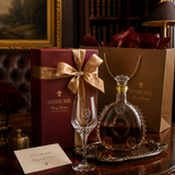 Louis XIII Bourbon cognac gift set on a dark wooden table, featuring a deep burgundy box with a gold ribbon, a brown paper shopping bag, a clear crystal coupe glass, and a decorated glass bottle of cognac on a silver tray.