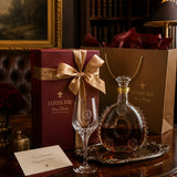 Louis XIII Bourbon cognac gift set on a dark wooden table, featuring a deep burgundy box with a gold ribbon, a brown paper shopping bag, a clear crystal coupe glass, and a decorated glass bottle of cognac on a silver tray.