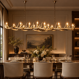 A large antler-style chandelier with multiple lit candle-style bulbs hangs over a formal dining table in an elegant, warm-toned dining room with upholstered chairs, floral arrangements, and wall art.