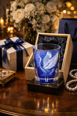 A cobalt blue etched glass tumbler featuring a white crane design, displayed in an open wooden gift box on a black base. The scene is set on a polished wooden table with a white gift box tied with a navy ribbon, pearl jewelry, and a blurred floral arrangement in the background.