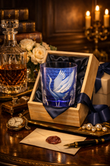 A blue etched glass tumbler featuring a white crane design, presented inside an open wooden gift box with dark blue satin lining. The scene is styled on a dark wooden table with a decanter of whiskey, a pocket watch, pearls, a pen, and a wax-sealed letter, under warm candlelight.