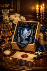 A blue etched glass tumbler with a white crane design, presented in a wooden gift box lined with dark blue satin. The box sits on a dark wooden table surrounded by a glass decanter, a glass of amber liquor, a pocket watch, pearls, a pen, and a wax-sealed envelope, with lit candles in the background.