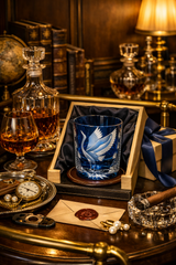 A blue etched crystal whiskey glass with a white swan design, presented in a wooden box on a dark table. The scene is styled with whiskey glasses, a decanter, books, a lamp, pearls, and a cigar in a warm, elegant, vintage-themed setting.