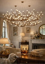 A large branch-shaped chandelier with many glowing bulbs hangs in a warm, elegantly furnished living room. The chandelier has a metallic gold/bronze frame with numerous light bulbs branching off it.