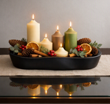 A styled holiday centerpiece in a black oval tray, featuring five lit pillar candles in cream, tan, and sage green, accented with pinecones, dried orange slices, cinnamon sticks, and red berries on a beige runner. A 'Made with AI' badge is visible in the top right corner.
