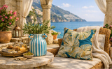 A styled outdoor patio scene with a wicker armchair draped in cream fabric, featuring a decorative throw pillow with a teal and gold palm leaf pattern. In the foreground, a round stone table holds a blue-and-white striped vase with greenery, candles, and small accessories.