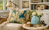 A styled living room scene with a beige armchair draped in cream fabric, featuring a prominent square throw pillow with a blue and gold palm leaf pattern. A matching blue and white striped ceramic vase with white flowers sits on a marble side table next to it, with a bookshelf and window in the background.