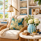 A styled living room scene with a beige armchair draped in cream fabric, featuring a prominent square throw pillow with a blue and gold palm leaf pattern. A matching blue and white striped ceramic vase with white flowers sits on a marble side table next to it, with a bookshelf and window in the background.