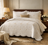 An elegant cream-colored quilted bedspread with scalloped edges and embroidered circular medallions, styled on a wooden bed frame in a classic bedroom setting with matching pillows and bedside tables.