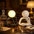 Two modern abstract desk lamps shaped like seated figures with round heads, shown on a wooden table in a warmly lit study. One lamp is matte black and glowing warm white; the other is matte white and unlit.