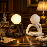 Two modern abstract desk lamps shaped like seated figures with round heads, shown on a wooden table in a warmly lit study. One lamp is matte black and glowing warm white; the other is matte white and unlit.