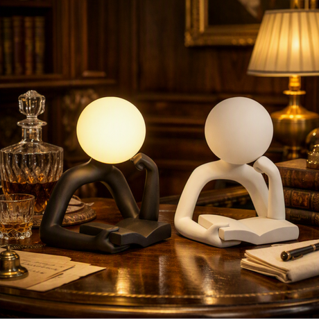 Two modern abstract desk lamps shaped like seated figures with round heads, shown on a wooden table in a warmly lit study. One lamp is matte black and glowing warm white; the other is matte white and unlit.