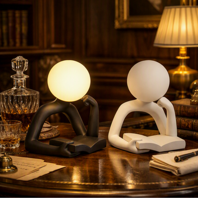 Two modern abstract desk lamps shaped like seated figures with round heads, shown on a wooden table in a warmly lit study. One lamp is matte black and glowing warm white; the other is matte white and unlit.