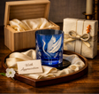 A blue glass candle holder with an etched white crane design, presented on a wooden tray with cream satin lining and a small card reading 'With Appreciation.' A wooden gift box and a wrapped gift box with a ribbon are visible in the background on a wooden table.