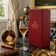Louis XIII cognac liqueur bottle, a filled coupe glass, and a deep red presentation box on a dark marble table in an elegant room setting.