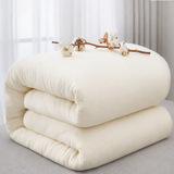 A folded off-white quilted mattress topper or comforter sits on a light gray floor in front of sheer white curtains. A decorative branch with cotton buds rests across the top.