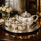 An ornate silver teapot and four matching cups with detailed dragon engravings, presented on a matching round tray on a dark wooden table. The background features a softly blurred warm interior with a vase of white roses and candles.