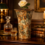 A tall, rustic ceramic vase with a mottled green, brown, and beige glaze sits on a dark wooden table in a warmly lit room. A single white rose is placed in the vase, alongside a decanter and a glass of amber liquid, with old books and a candle in the background.