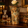 A tall, rustic ceramic vase with a mottled green, brown, and beige glaze sits on a dark wooden table in a warmly lit room. A single white rose is placed in the vase, alongside a decanter and a glass of amber liquid, with old books and a candle in the background.