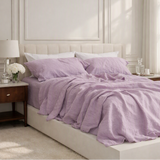 A bed dressed in light lavender bedding, including a duvet cover and two matching pillows, styled in a bright, modern bedroom with a cream upholstered headboard, wooden nightstands, and a window with sheer curtains.
