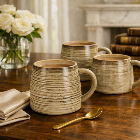 Three rustic ceramic mugs with a textured horizontal stripe pattern in beige and brown tones, arranged on a wooden table with a gold spoon, a vase of white roses, and books in the background.
