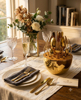 A styled dining table setting featuring a reflective gold bowl-shaped cutlery holder filled with gold-toned flatware, alongside matching gold cutlery on plates with dark gray napkins, crystal glasses, a floral centerpiece, and a card reading AURUM.