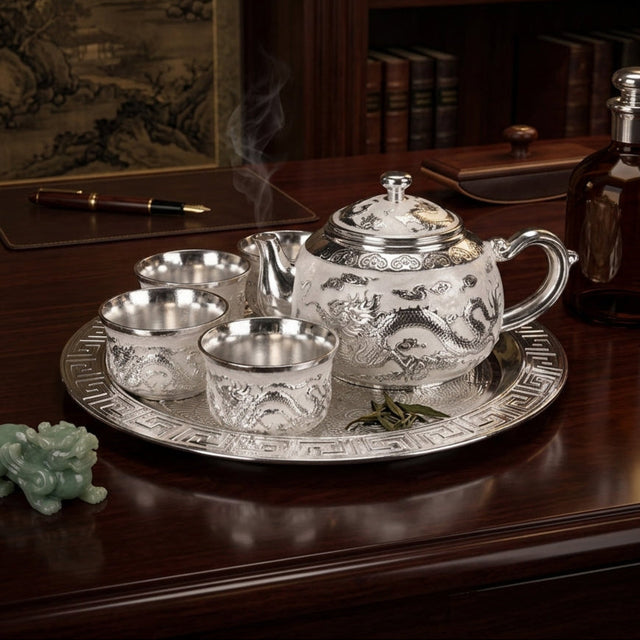 A silver teapot and four matching teacups with dragon engravings, set on a round silver tray on a dark wooden table. Steam rises from the teapot, and a small green jade figurine sits nearby.