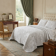 A light gray floral jacquard quilted bedspread set with matching pillowcases on a bed in a classic, elegant bedroom with green drapes, a wooden desk, and parquet flooring.